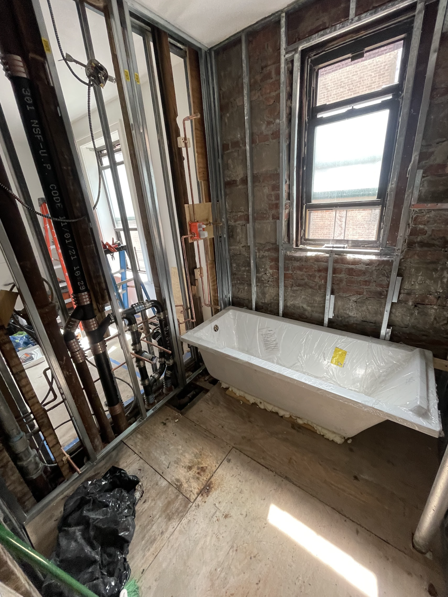 New bathtub positioned during bathroom rough-in in a Bushwick pre-war tenement, exposed brick walls and wood subfloor visible during gut renovation