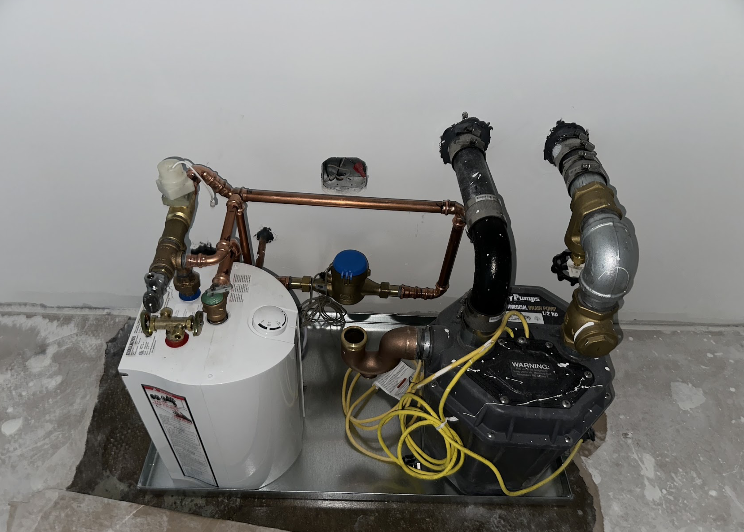 Prefabricated water heater manifold assembly with white expansion tank, circulator pump, brass fittings, and copper piping installed in a Bushwick multi-family building mechanical room