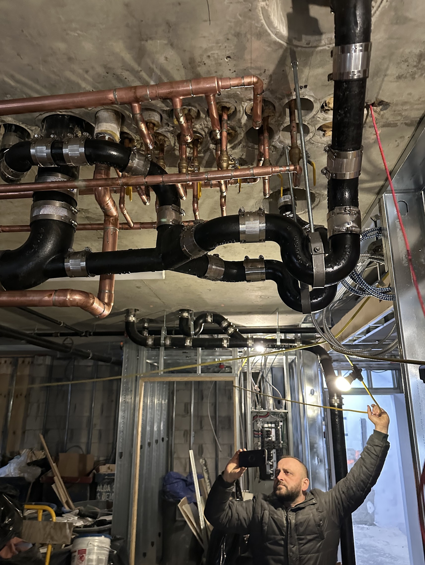 Yellow PEX tubing connected to copper supply lines with expansion tank, brass ball valves, and manifold components for a radiant floor heating rough-in in a Ridgewood two-family home